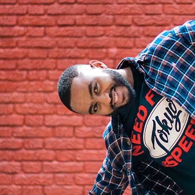 Guy with black shirt and blue plaid shirt in front of a red brick wall.