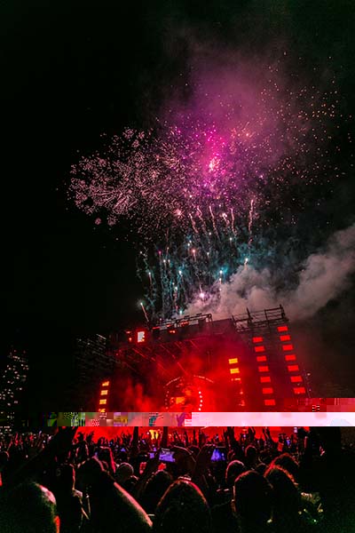 Dark outdoor concert with red stage lights, a large crowed and fireworks going off.