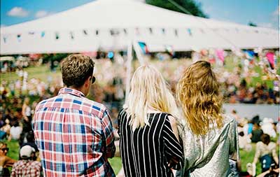a man and two woment sitting with thier backs facing the camera looking out into a carnival tent and field full of people.