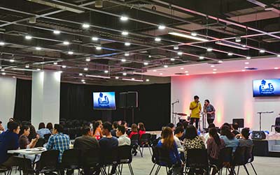 a room with circular tables filled with guests and a small white stage with two speakers addressing the audience.